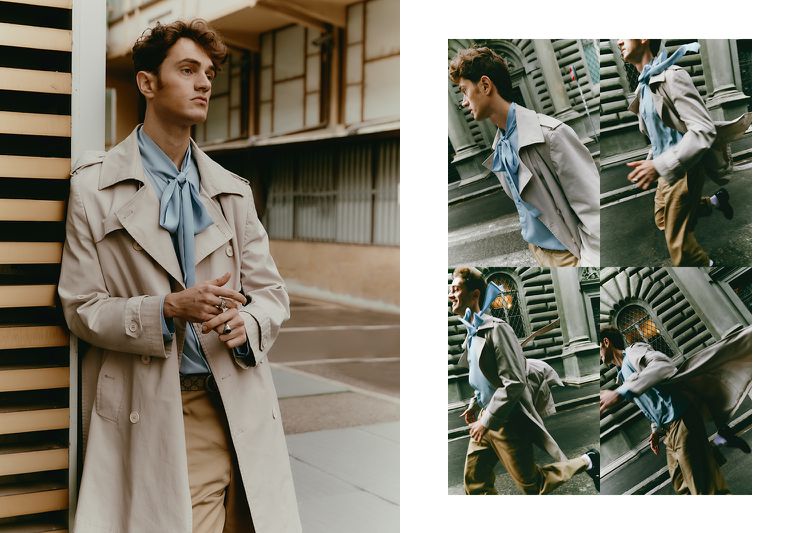 A young adult male in a trench coat and light blue shirt stands in an urban setting with beige and tan tones, captured with natural light.