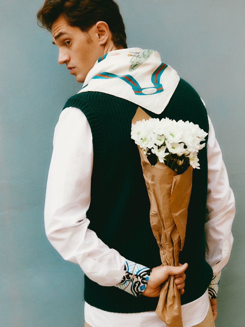 Young man in green sweater vest, white shirt, and patterned scarf hides bouquet of flowers behind his back against a blue wall.