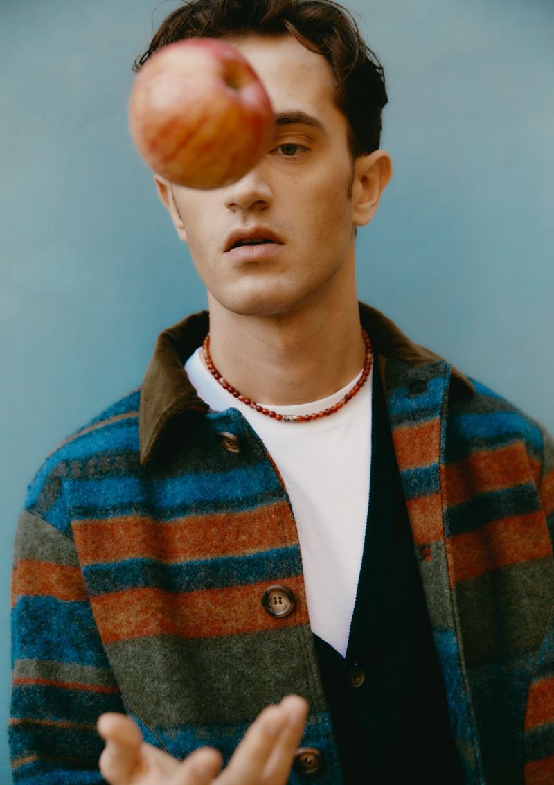 Young man wearing a striped shirt and necklace looks at a levitating apple in front of a teal backdrop in a studio portrait.
