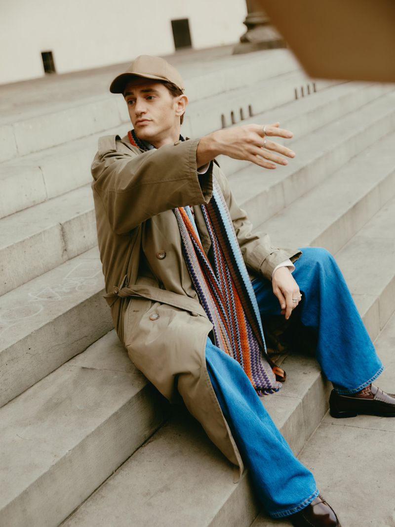 An adult male sits on concrete steps wearing a trench coat, scarf, jeans, and a baseball cap while gesturing with his hand.