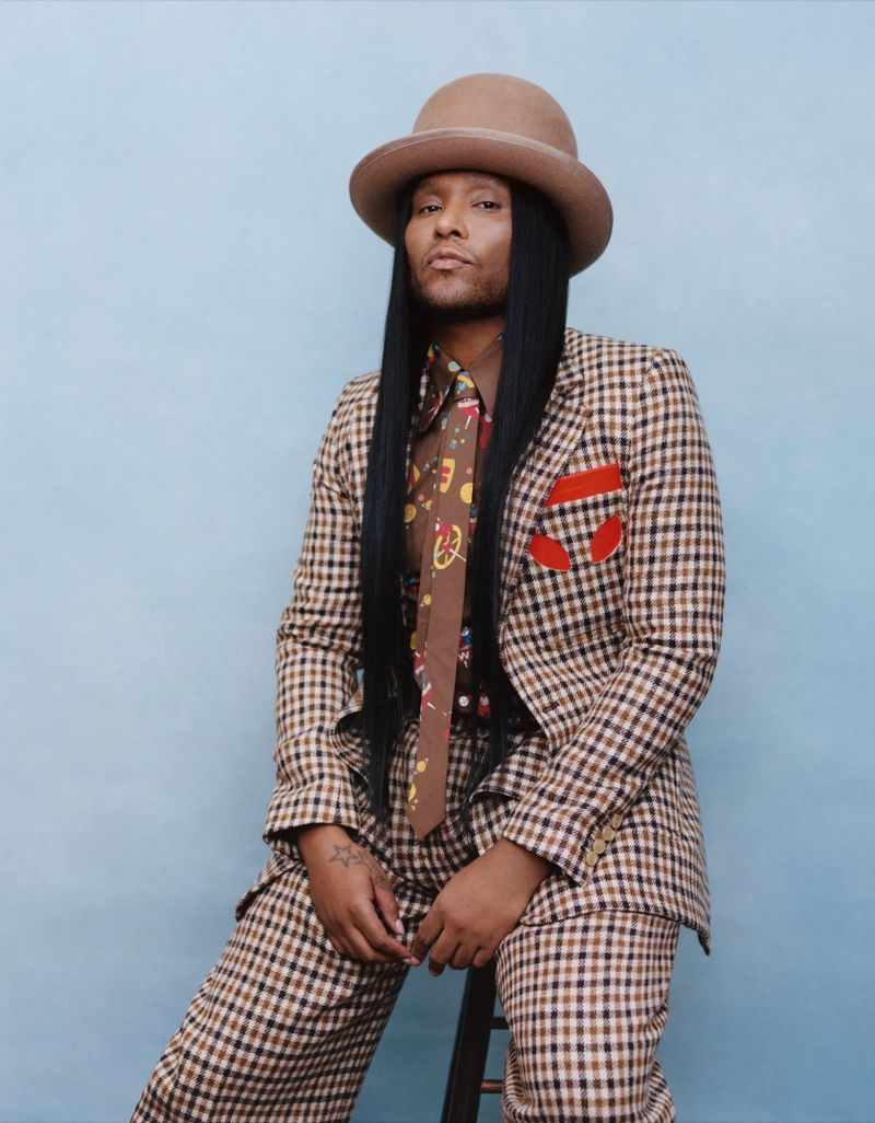 Person posing in a brown plaid suit and hat against a light blue background, full view, studio lighting, fashion photography.