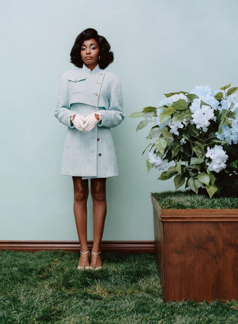Elegant Black woman in a light blue tweed suit, posing near a hydrangea bush and green lawn against a light blue background.
