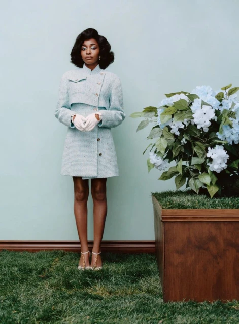 Elegant Black woman in a light blue tweed suit, posing near a hydrangea bush and green lawn against a light blue background.