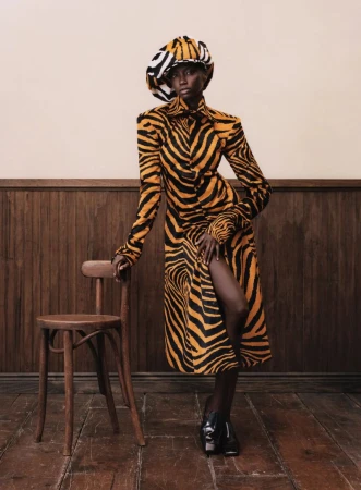 Model in zebra-print coat and matching hat standing beside a wooden chair against a wood-paneled wall