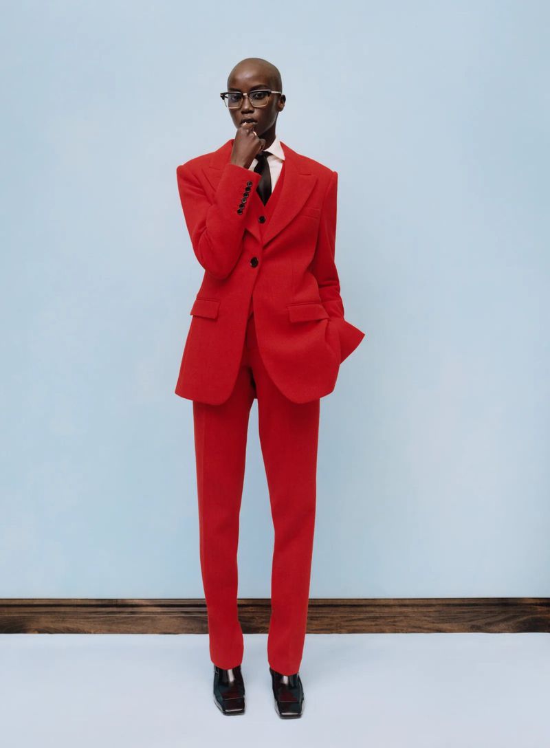 Full-length portrait of a Black female model in a bright red suit, black tie, and glasses, standing in a photo studio against a light blue backdrop.