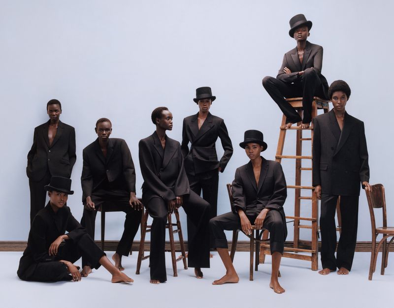 Group of dark-skinned fashion models wearing black suits and hats, seated and standing in front of a pale blue background.