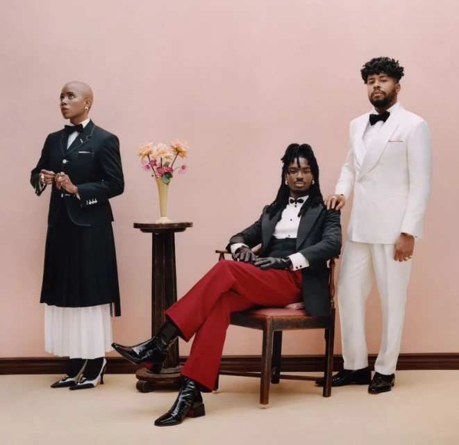 Three people in formal wear pose in front of a pale pink background. One person sits in a chair, another stands, and a vase of flowers sits on a table.