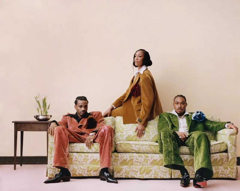 Three people in vibrant velvet suits pose on a floral couch with a vintage side table and tulips in a stylish indoor setting.