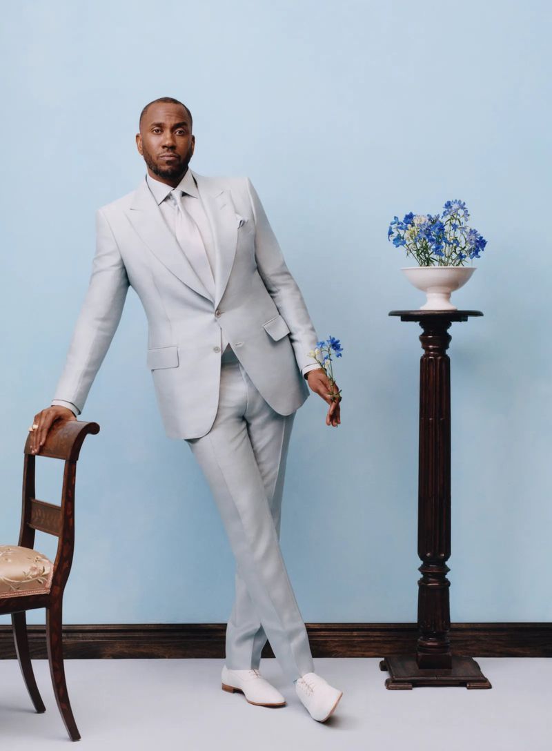 Man wearing a light blue suit standing against a pale blue backdrop leaning on a wooden chair