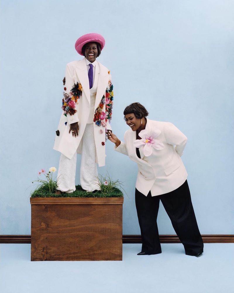 Two women in embellished formal wear pose in a studio against a light blue backdrop. One woman stands on a wooden platform.