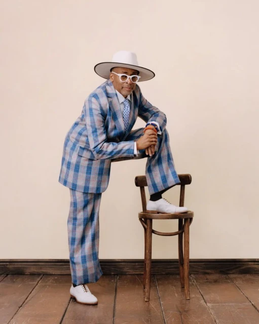 Adult male in a blue plaid suit, hat, and white shoes, posing on a wooden chair in a neutral setting with a wooden floor.