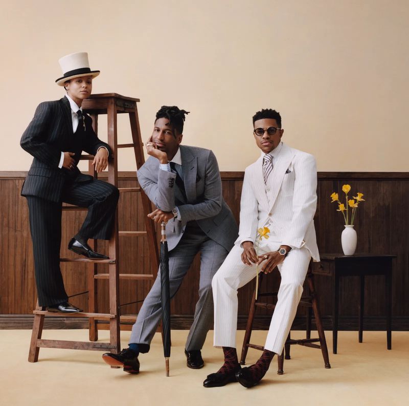 Three fashionable Black men in formal suits pose in an indoor setting with flowers. One stands on a ladder and wears a top hat.