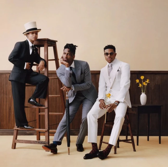 Three fashionable Black men in formal suits pose in an indoor setting with flowers. One stands on a ladder and wears a top hat.