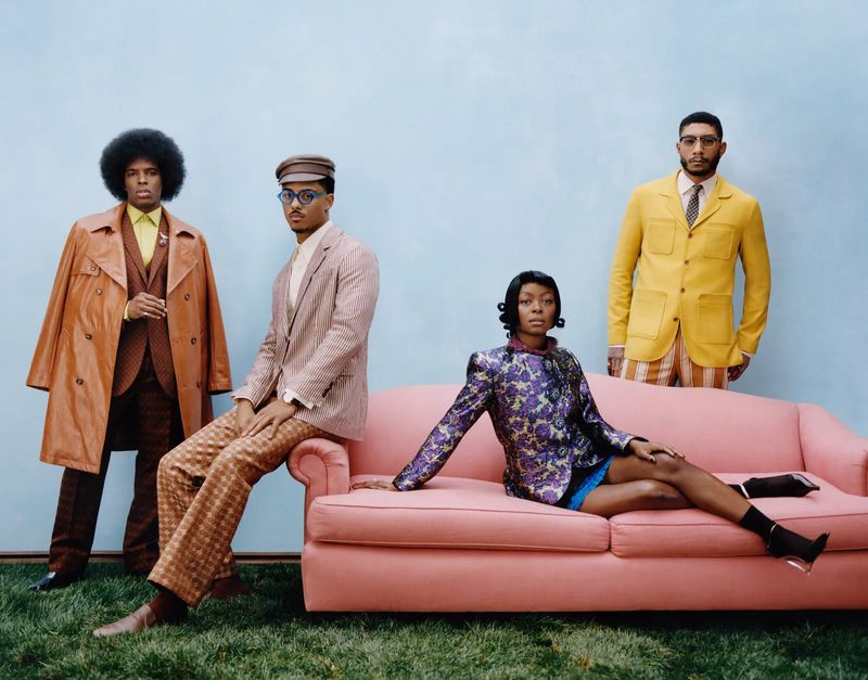 Four stylish people pose against a blue backdrop. One woman sits on a pink couch, while three men stand or sit in coordinated vintage outfits.
