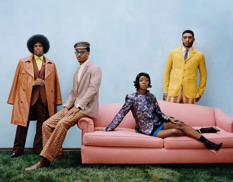 Four stylish people pose against a blue backdrop. One woman sits on a pink couch, while three men stand or sit in coordinated vintage outfits.