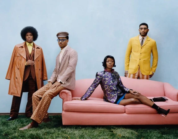 Four stylish people pose against a blue backdrop. One woman sits on a pink couch, while three men stand or sit in coordinated vintage outfits.