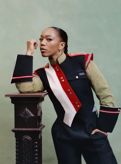 Headshot of a woman with her hand near her face, wearing a navy and red vest, posing near a dark column against a light green background.