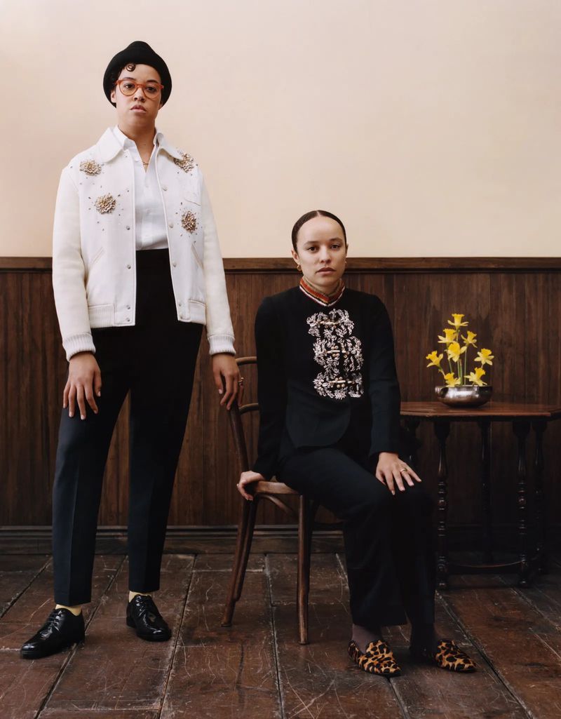 Two women in formal attire pose indoors. One is standing in a white jacket and the other is sitting wearing a black jacket.
