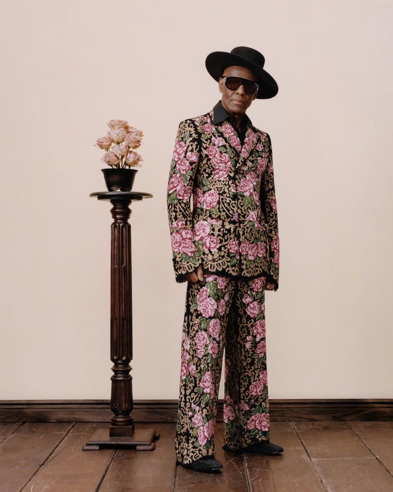 Man in floral suit stands next to pedestal with roses, wearing sunglasses and hat, against a neutral wall and wooden floor.