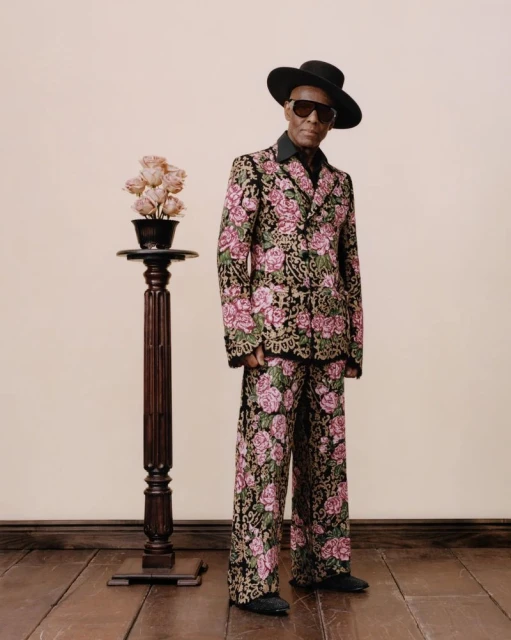 Man in floral suit stands next to pedestal with roses, wearing sunglasses and hat, against a neutral wall and wooden floor.