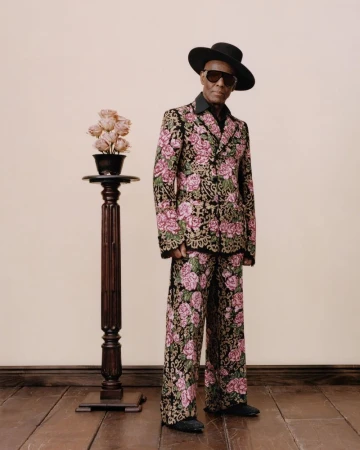 Man in floral suit stands next to pedestal with roses, wearing sunglasses and hat, against a neutral wall and wooden floor.
