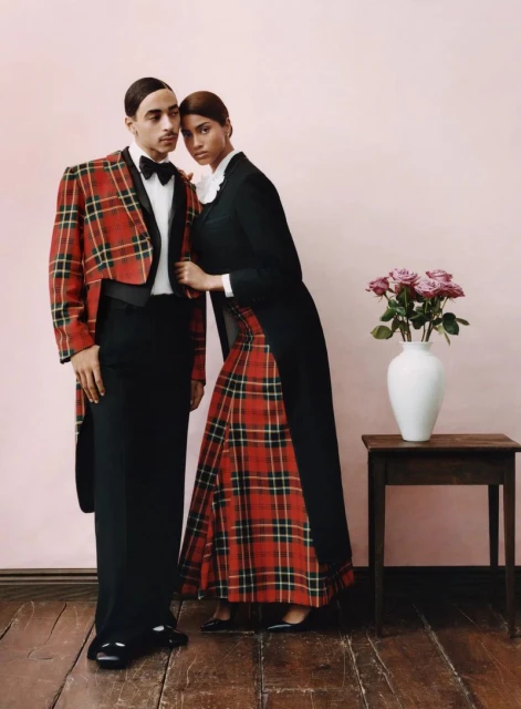 A smartly dressed couple poses in a studio setting. The man wears a red plaid jacket and the woman a matching plaid skirt.