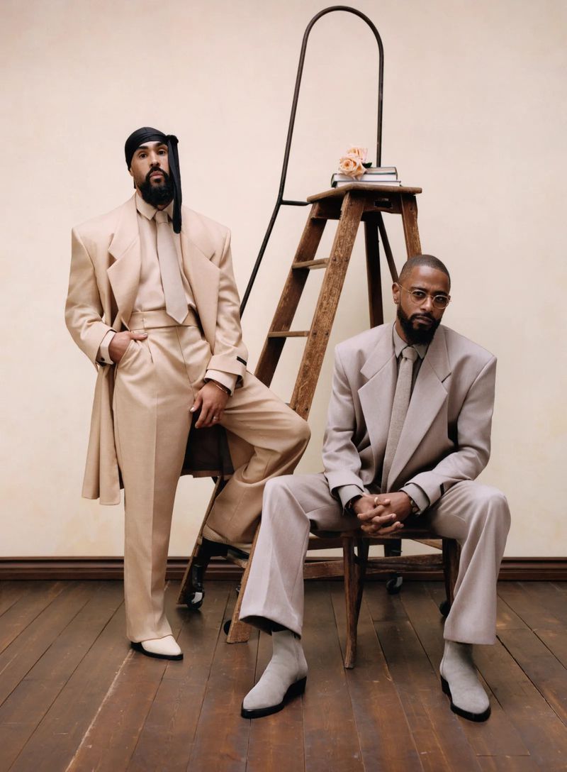Two adult males in tan suits pose indoors near a wooden ladder holding books and light pink flowers against a cream background.