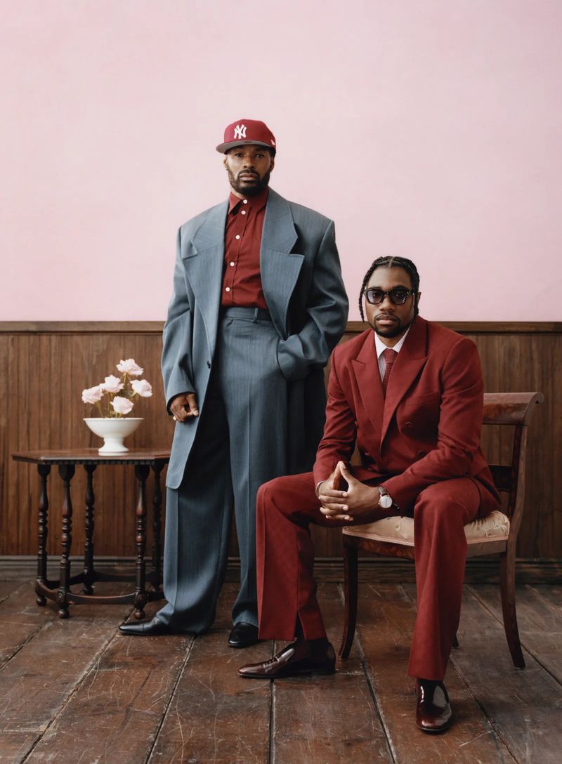 Two Black men pose indoors: one stands in an oversized grey suit with a red baseball cap; the other sits in a red suit and glasses.