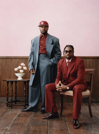 Two Black men pose indoors: one stands in an oversized grey suit with a red baseball cap; the other sits in a red suit and glasses.