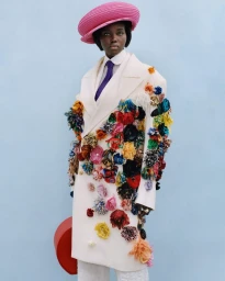 Model wearing a white floral-apprliqued coat and pink beret standing against a light blue background for American Vogue Dandyism