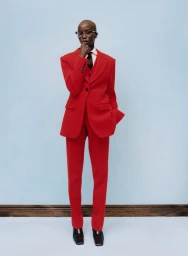 Model in a red suit wearing glasses and a tie standing against a light blue backdrop for American Vogue Dandyism project