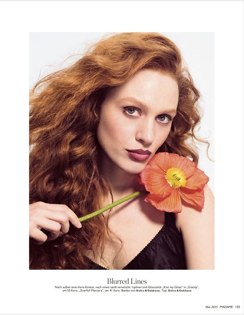 Close-up shot of an adult woman with auburn hair and black top holding a poppy flower against a neutral background in a studio setting.