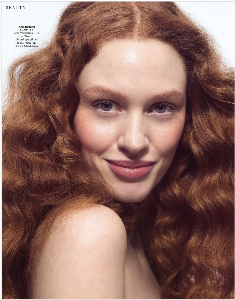 Close-up portrait of a fair-skinned woman with long, wavy auburn hair, light blue eyes, and a faint smile against a white background.