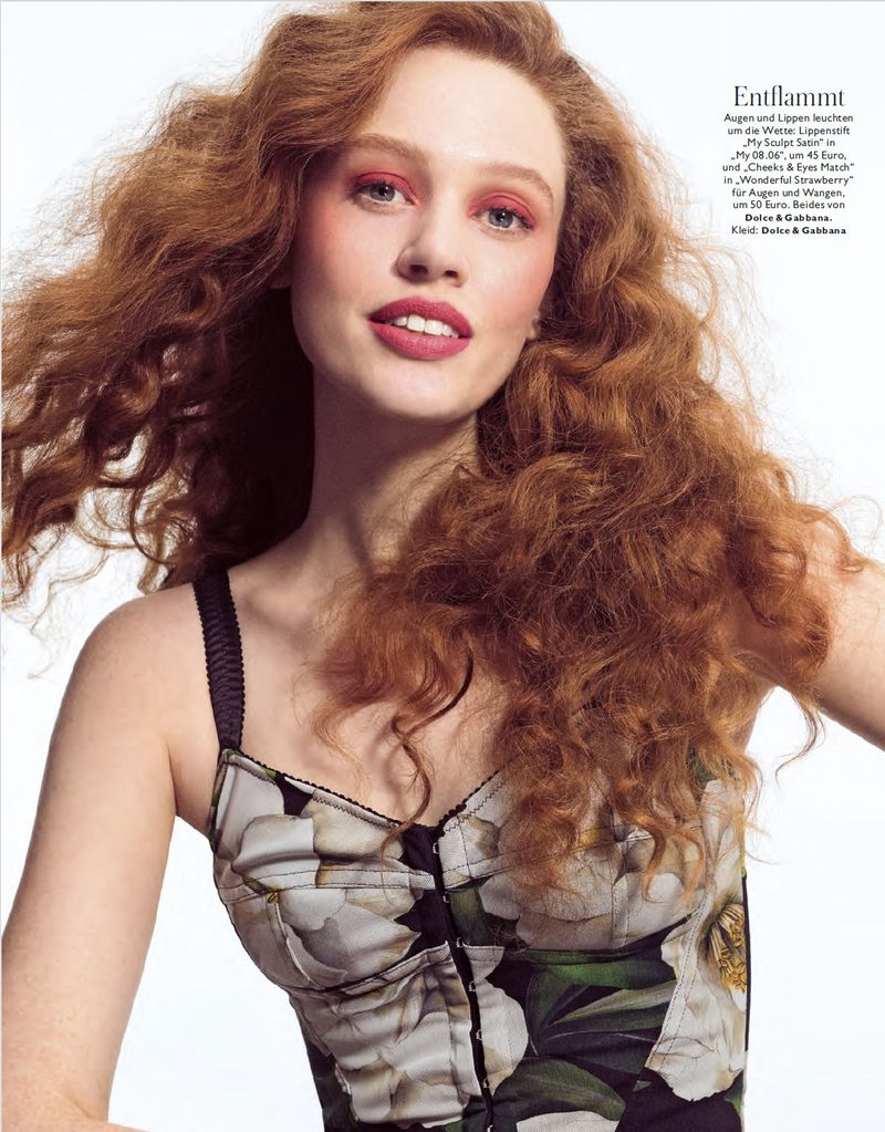 A red-haired woman smiles softly in a close-up fashion portrait, wearing a floral bustier top against a white background in a studio setting.