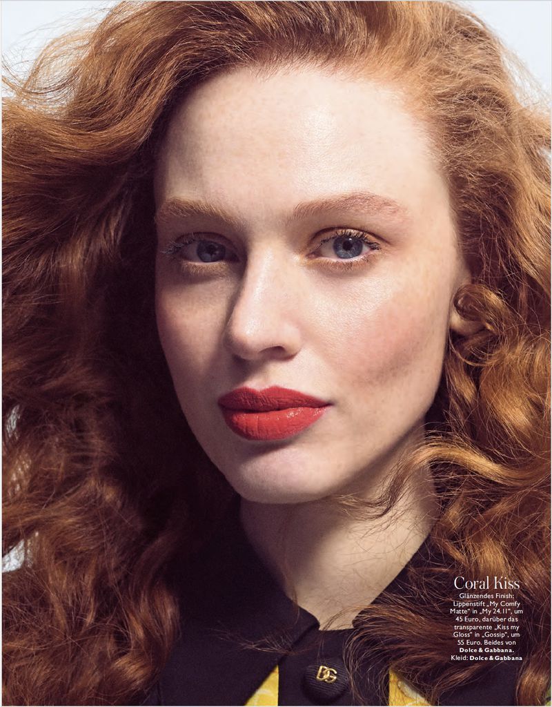 Close-up beauty shot of a red-haired, fair-skinned young adult woman with blue eyes and coral lipstick wearing a dark blouse.