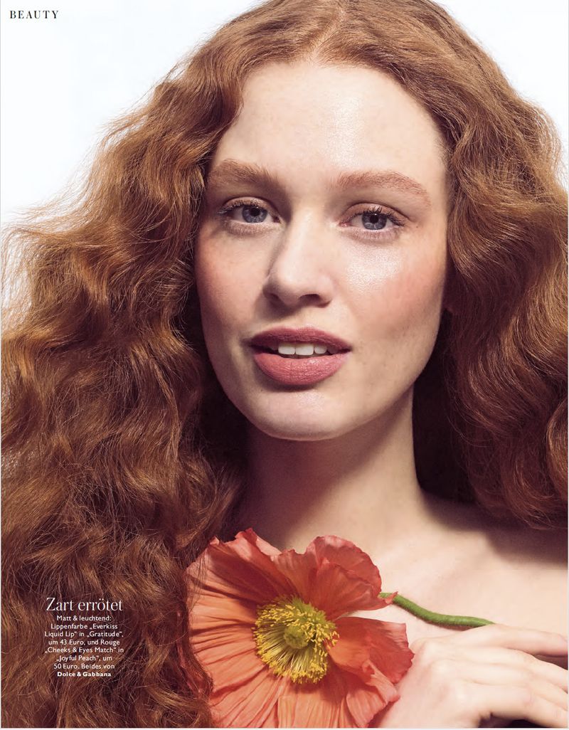 Close-up studio shot of a red-haired adult woman with light skin, wearing makeup and holding an orange poppy in a beauty context.