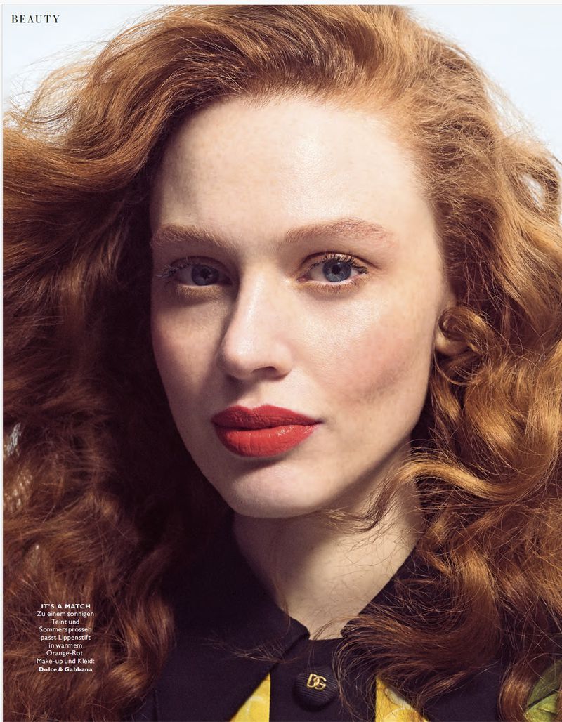 Close-up beauty shot of a red-haired model with bright red lips wearing a black and yellow patterned shirt with a DG logo button.
