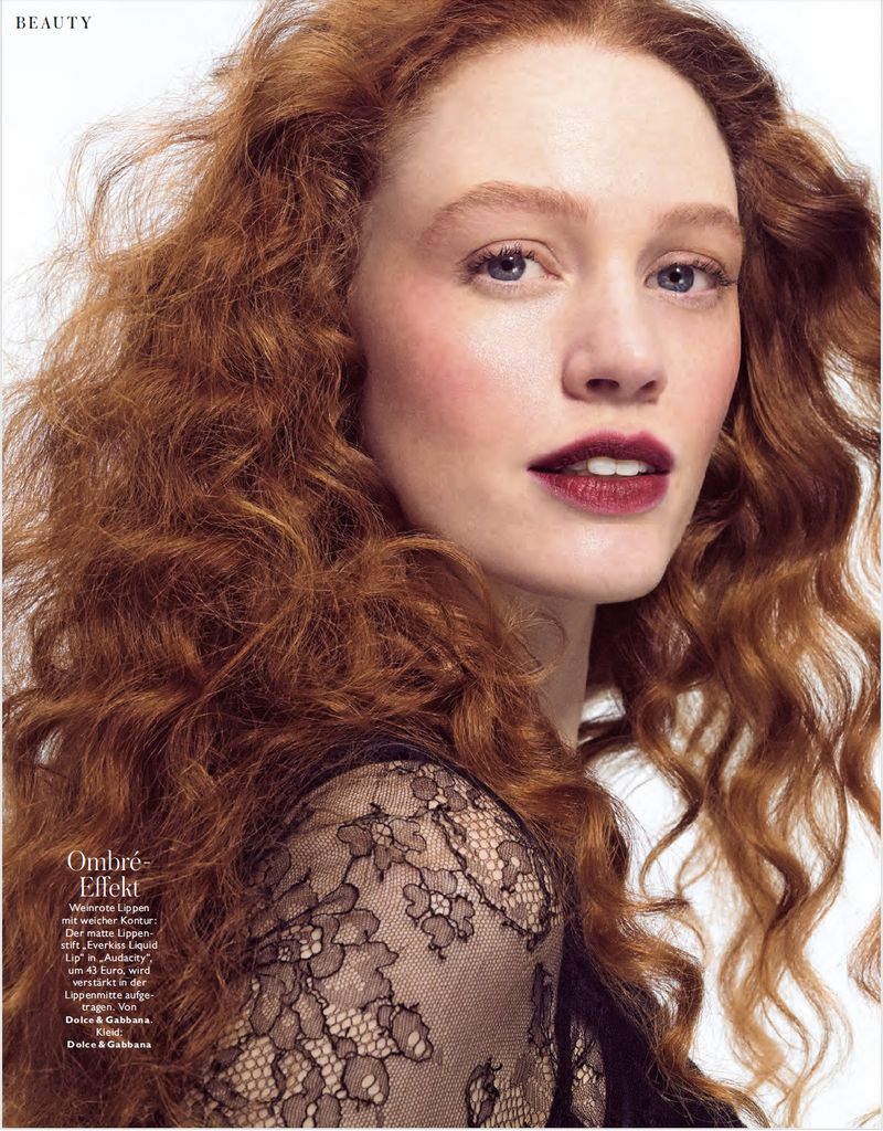 Close-up beauty portrait of a woman with curly red hair and dark burgundy lipstick wearing black lace against a white background