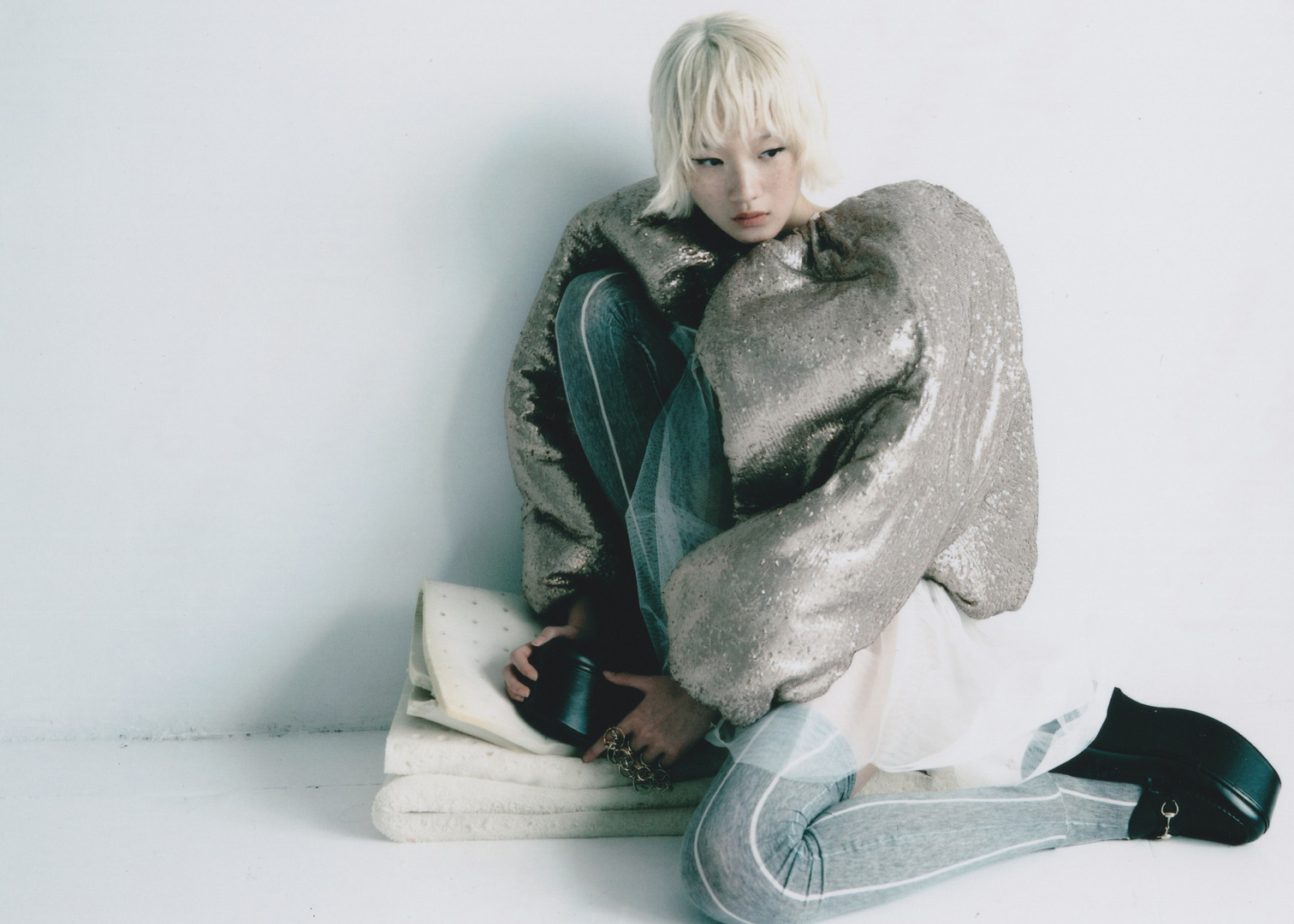 A young person with a silver sequin jacket poses in a studio. They wear dark platform shoes, grey pants, and a white top, set against a white backdrop.