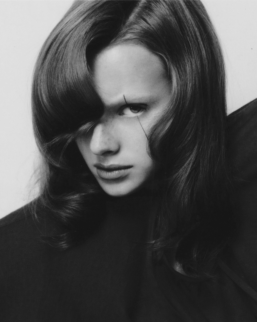 Dramatic black-and-white portrait of a young adult female with striking hair and freckles, wearing a dark top, against a neutral background.