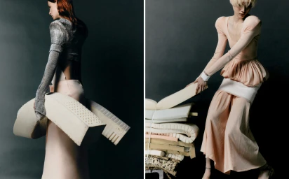 Two models posing in sculptural fashion garments while lifting sections of foam mattresses against a dark grey backdrop.