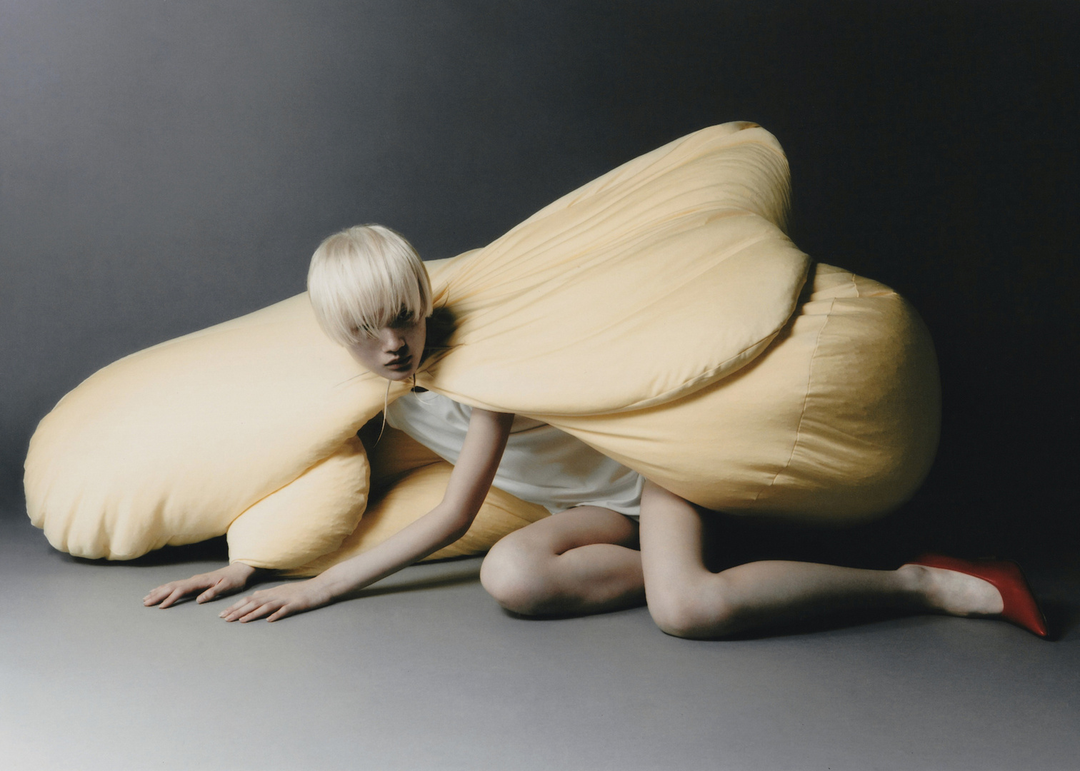 Blonde hair model wearing a large, soft yellow structure and red heels. Studio shot against a dark grey background.