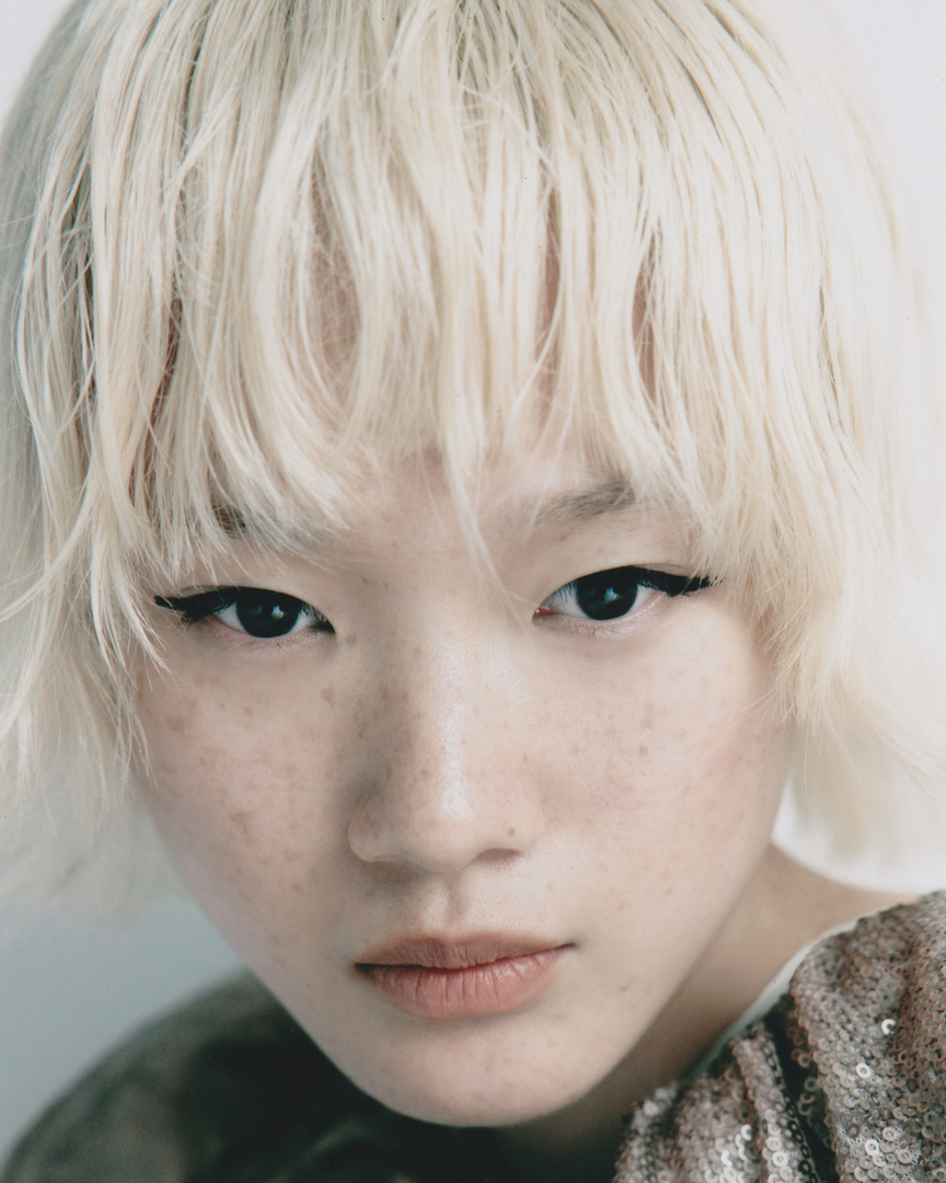 Close-up portrait of an Asian woman with platinum blonde short hair, freckles, and neutral facial expression, wearing a sequined top.