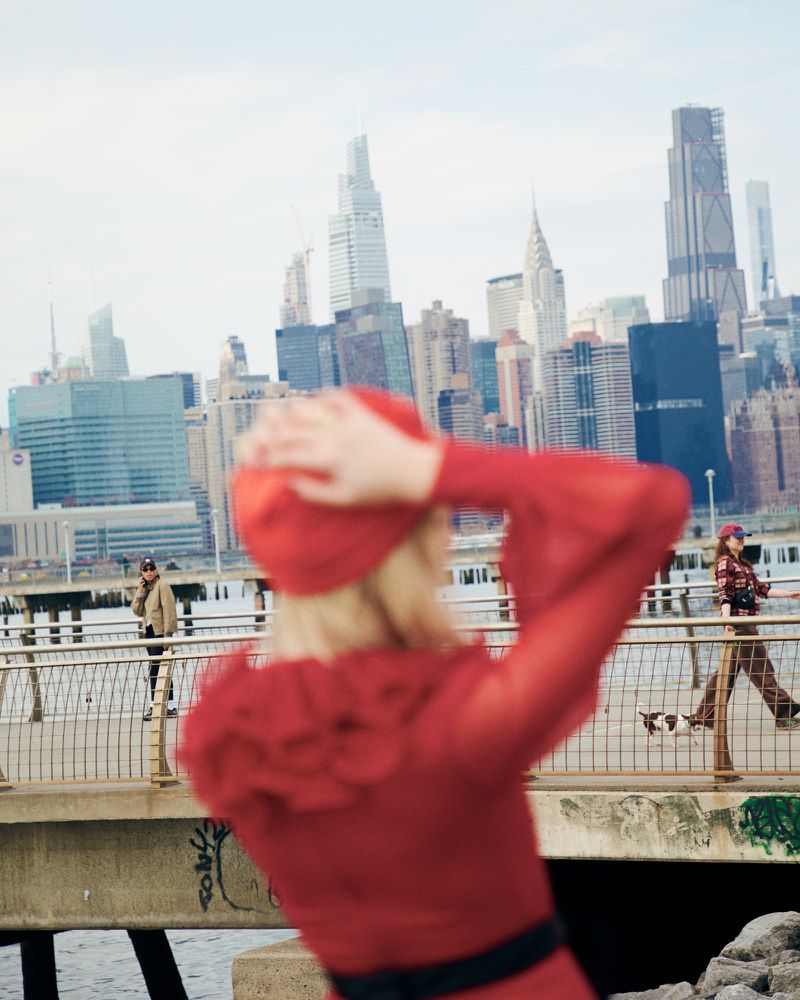 Out-of-focus woman in red dress and hat poses on waterfront with NYC skyline backdrop, joined by other figures and a dog.