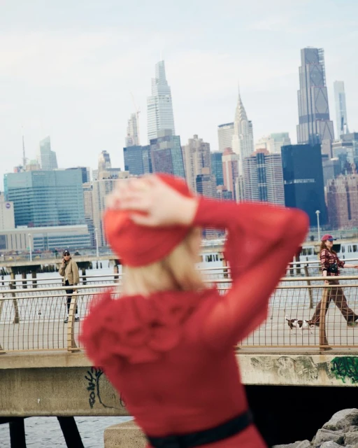 Out-of-focus woman in red dress and hat poses on waterfront with NYC skyline backdrop, joined by other figures and a dog.