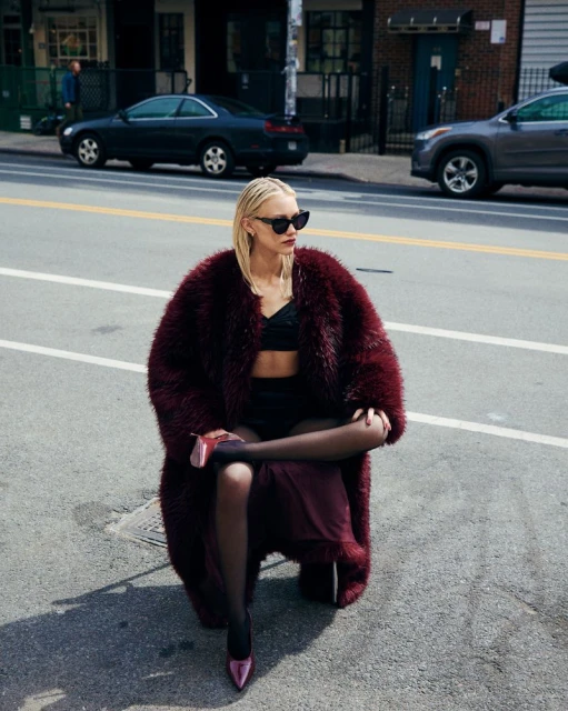 A blonde woman in a burgundy fur coat and black sunglasses sits on an urban street with cars and buildings in the background.