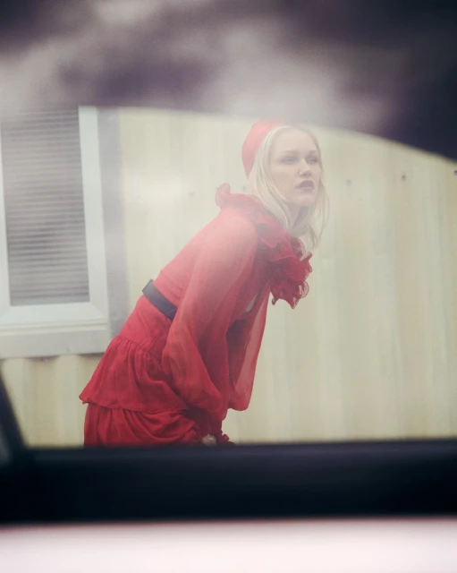 Woman in red dress and hat looks away from car window, with background building and cloudy sky visible. Fashion editorial style.