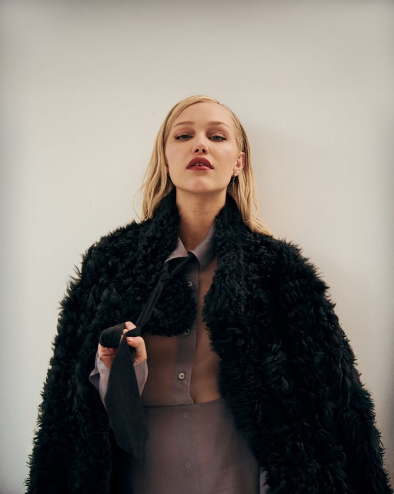 Portrait of a blonde woman wearing a shearling coat and holding a tie, posing indoors against a neutral wall in soft, diffused light.