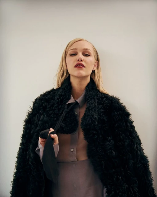 Portrait of a blonde woman wearing a shearling coat and holding a tie, posing indoors against a neutral wall in soft, diffused light.