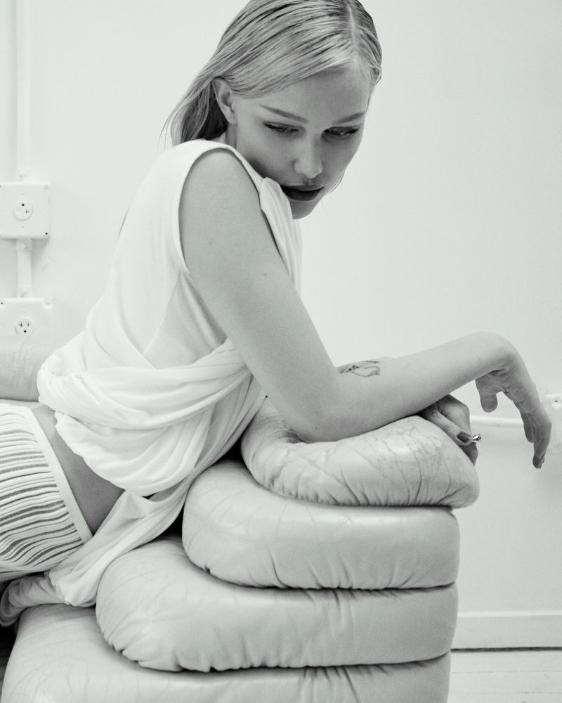 Monochrome image of a blonde woman in a white top and striped bottoms reclining on a stacked cushion, with pale skin and dark lipstick.